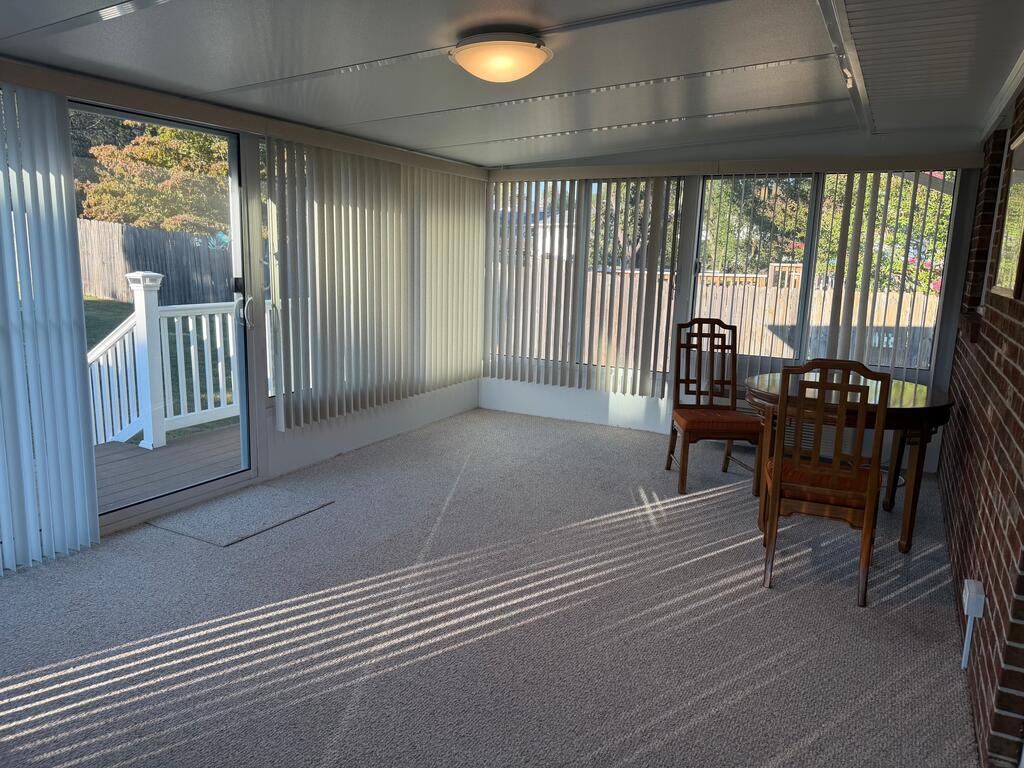Enclosed sunroom with vertical blinds, carpet, and a wooden table and chairs beside a deck door.