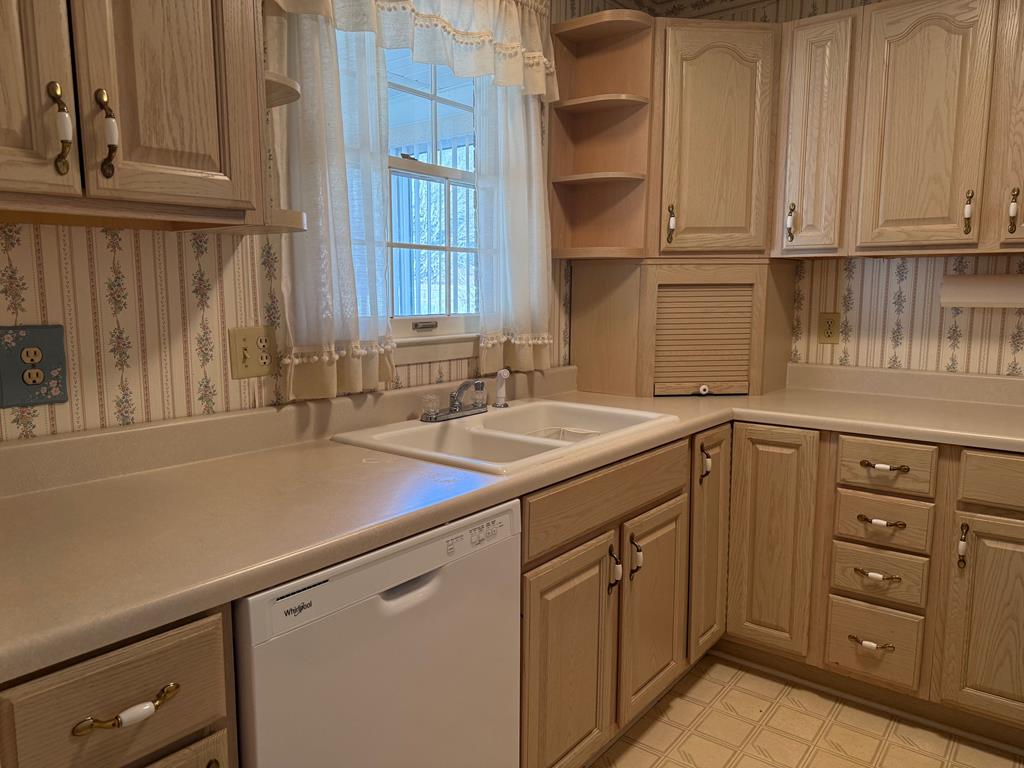 Kitchen with light wood cabinetry, a double-basin sink beneath a curtained window, and a white dishwasher.