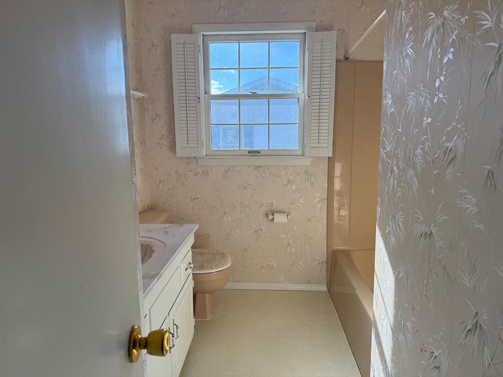 Bathroom with pale floral wallpaper, a shuttered window, white vanity, toilet, and bathtub.