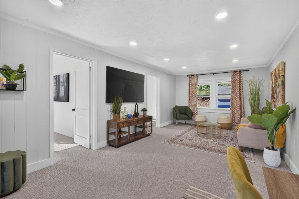 Staged living room with a wall-mounted television, patterned rug, and seating by the windows.
