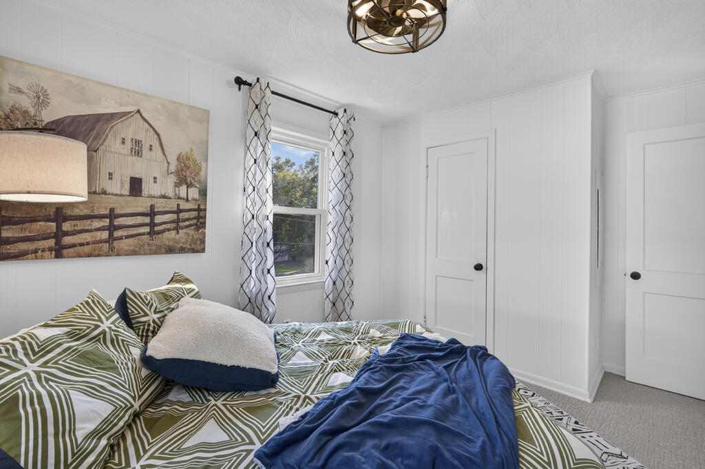 Bedroom with green patterned bedding, a blue throw, farmhouse artwork, and a window with patterned curtains.