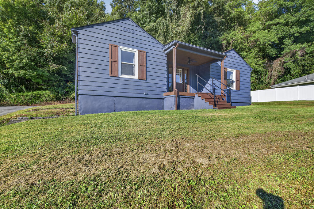 Blue-sided house with a covered porch stands above a sloping lawn, backed by mature trees.