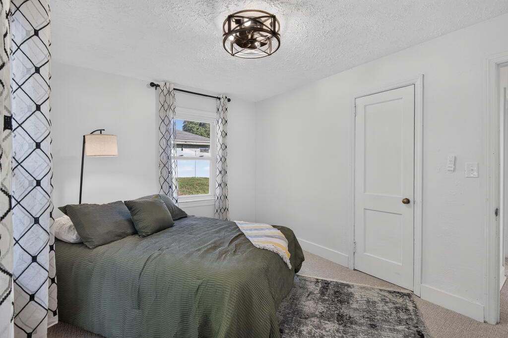 Bedroom with muted green bedding, patterned curtains, and a gray area rug.