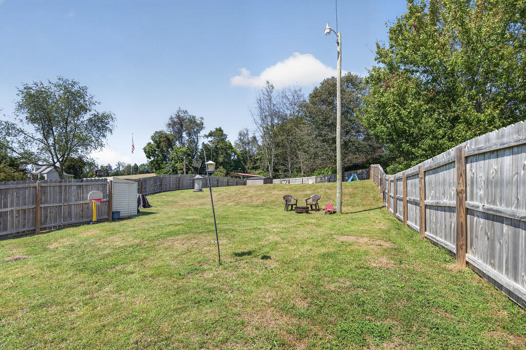 Fenced backyard with a broad lawn, basketball hoop, play structure, and chairs around a fire pit.