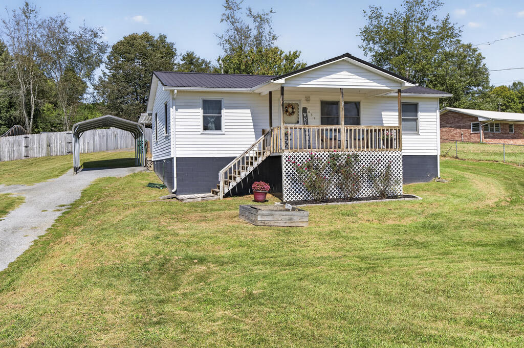 White single-story home with a covered porch, dark metal roof, and gravel driveway beside a carport.