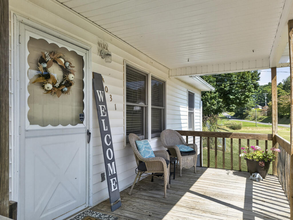 Covered front porch with a wreath-decorated white door, wicker chairs, and potted flowers overlooking a green yard.