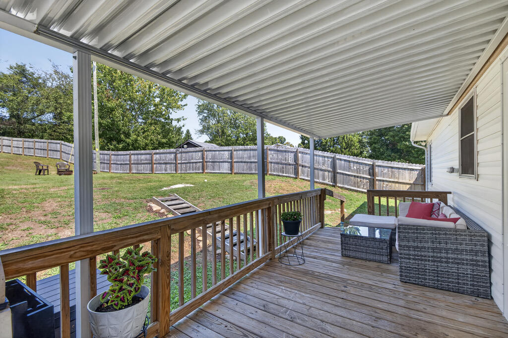 Covered wooden deck with wicker seating and potted plants overlooking a fenced backyard.
