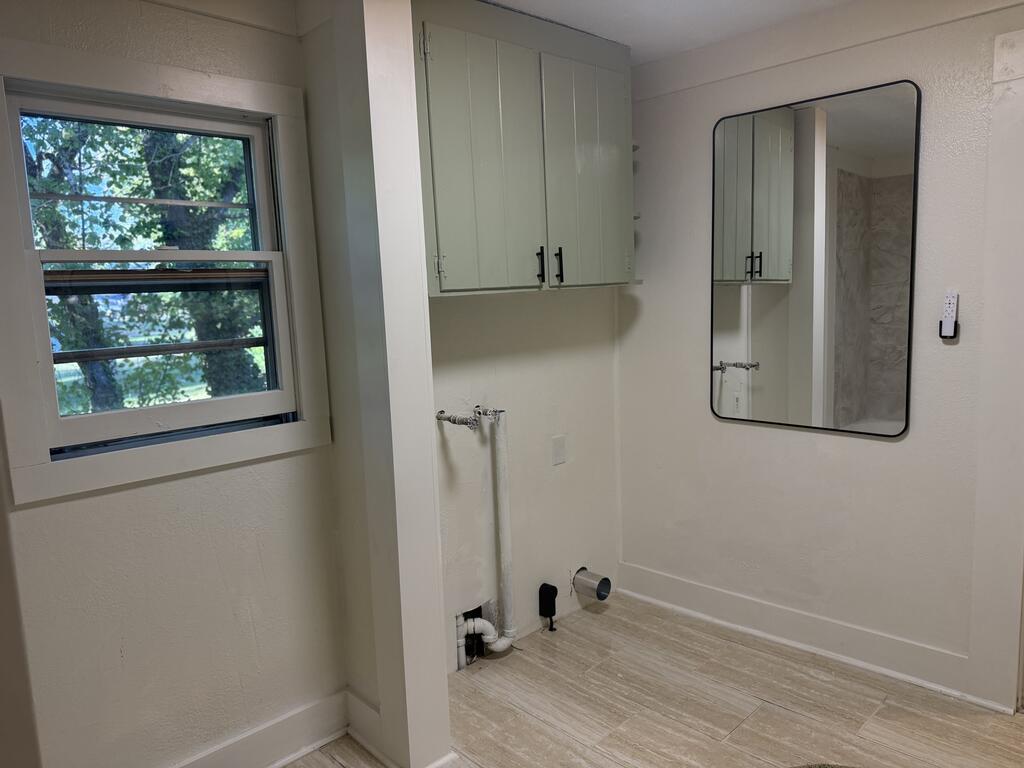 Bathroom with pale wall cabinets, a large mirror, exposed plumbing, and a window overlooking trees.