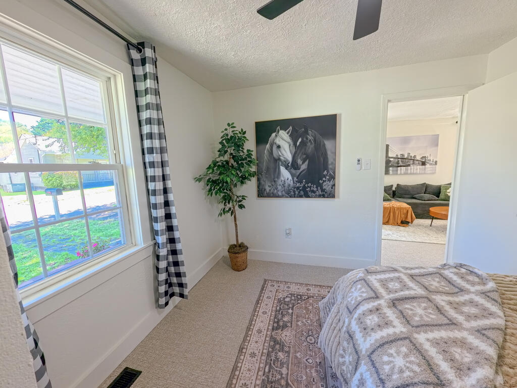Furnished bedroom with check curtains, horse artwork, and an open doorway to the living room.