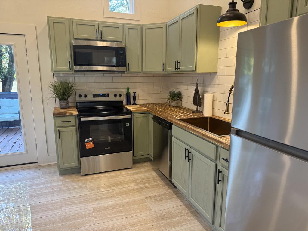 Kitchen with sage green cabinets, stainless steel appliances, butcher-block counters, and a dark farmhouse sink.