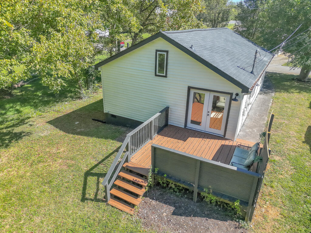 Rear of a white-sided house with a raised wooden deck and stairs beneath mature trees.
