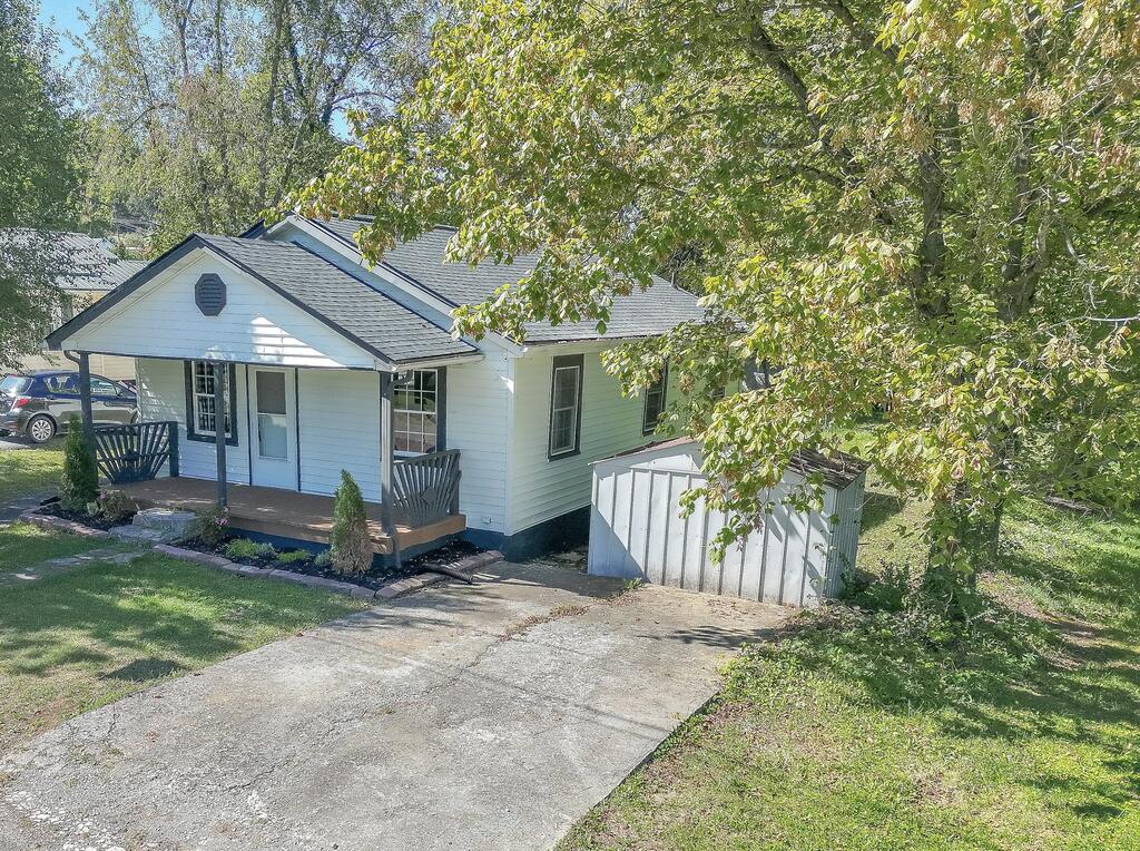 White single-story house with a covered porch, concrete driveway, and leafy trees.