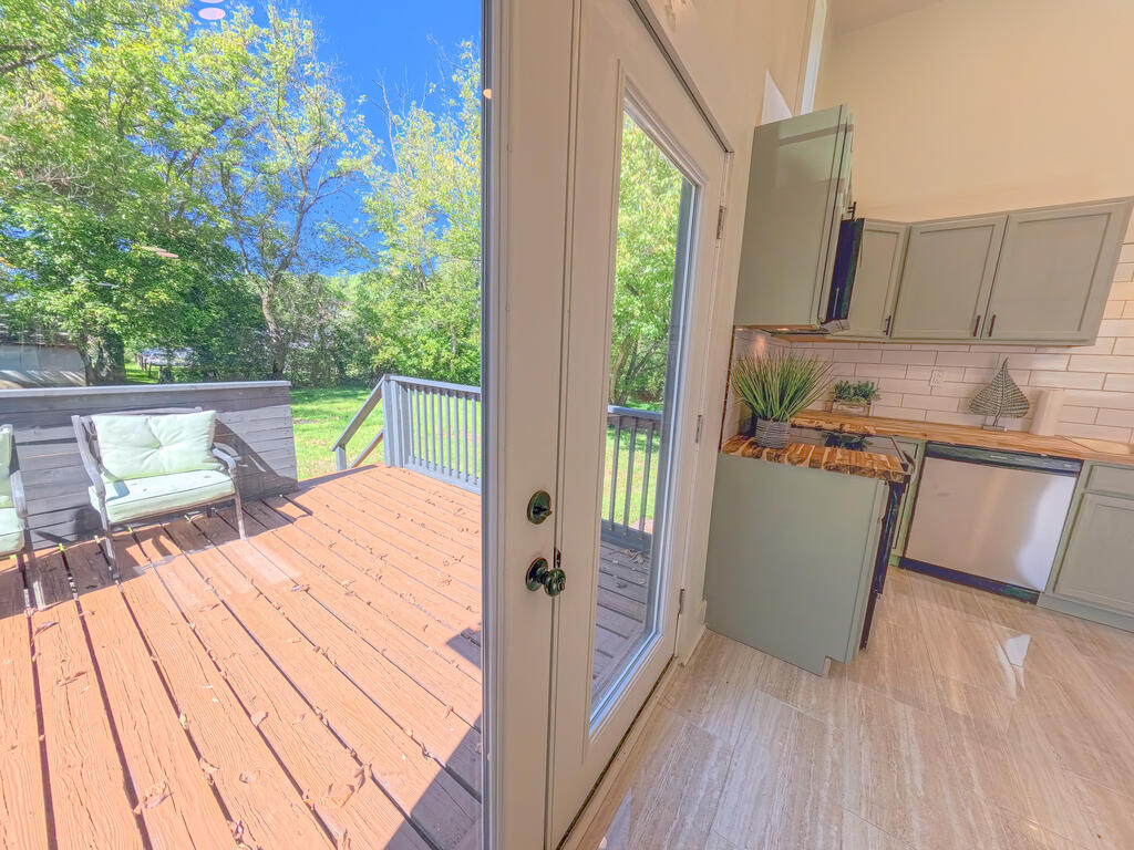 Kitchen opening onto a sunny wooden deck with a white cushioned chair and leafy trees.