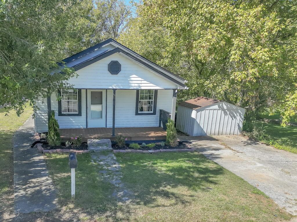 Small white house with dark blue trim, a covered porch, and a detached shed under leafy trees.