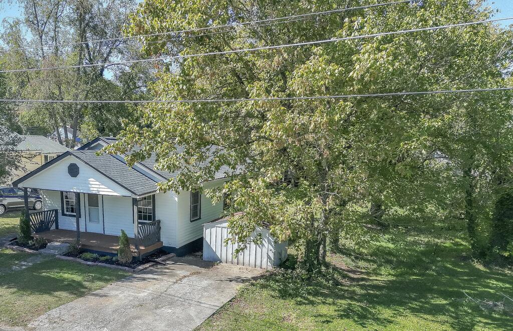Small white house with a covered porch, concrete driveway, and mature trees shading the yard.