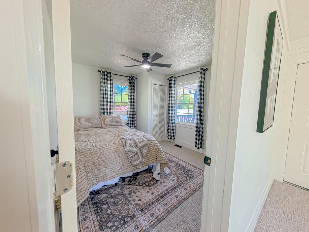 Bedroom with a beige quilted bed, patterned rug, ceiling fan, and checkered curtains.