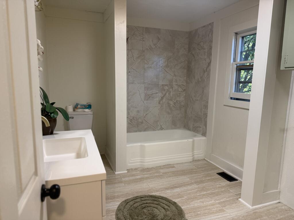 Bathroom with a white vanity, marble-pattern tub surround, window, and round rug.