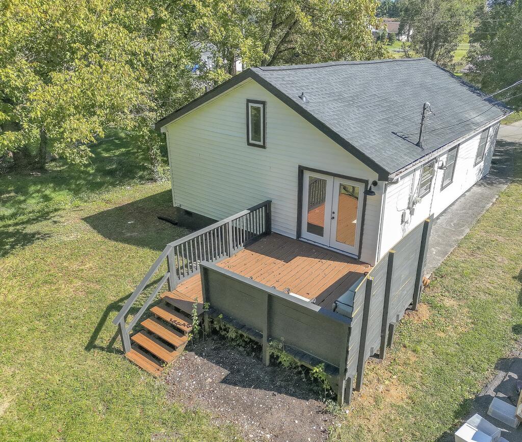 Rear of a light-colored house with a raised wooden deck and stairs, surrounded by a leafy lawn.