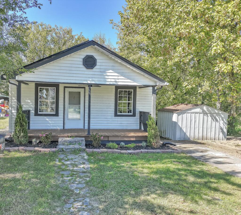 Small light-sided house with a covered porch, stone walkway, and metal shed beside it.