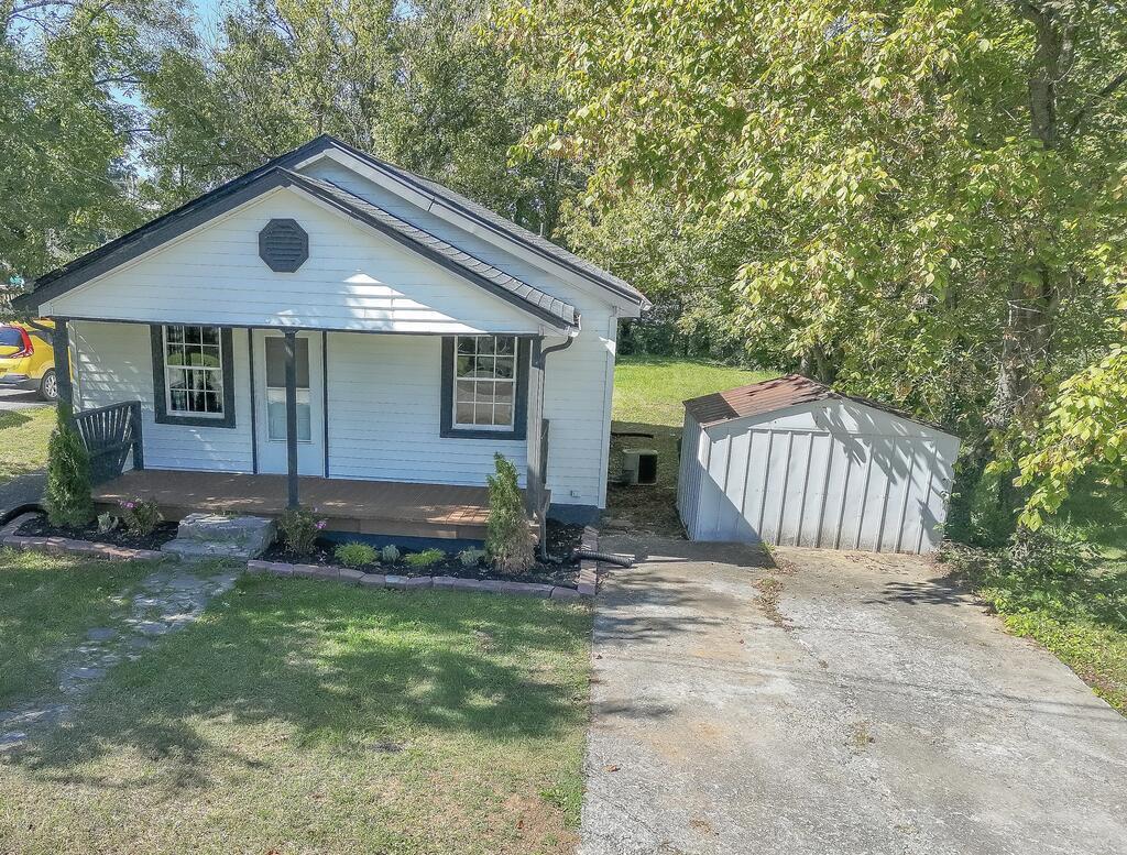 Small white house with a covered porch, concrete driveway, and detached shed among mature trees.