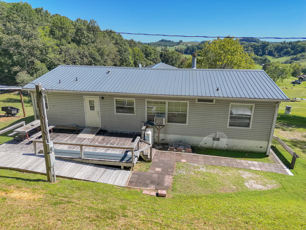 Rear of a gray-sided home with a metal roof, wooden deck, and paved patio amid wooded hills.
