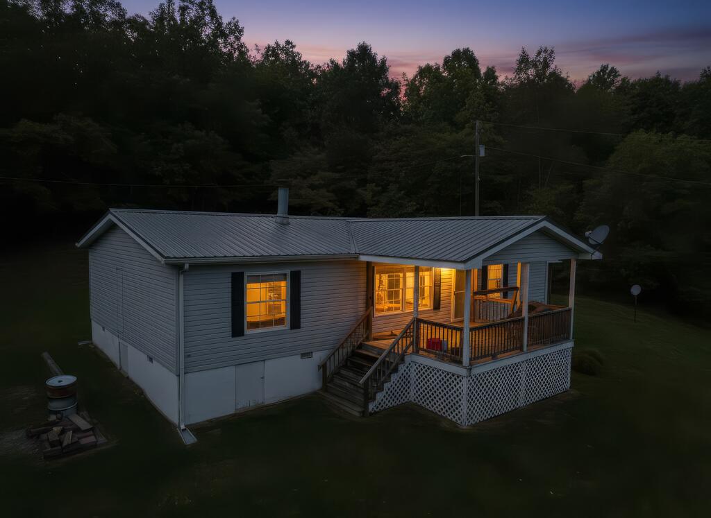 Gray-sided home with a metal roof and illuminated covered porch at dusk, bordered by trees.