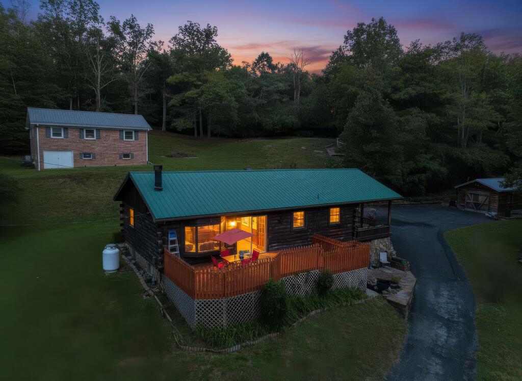 Log cabin with a green metal roof and warmly lit windows beside a raised deck at dusk.