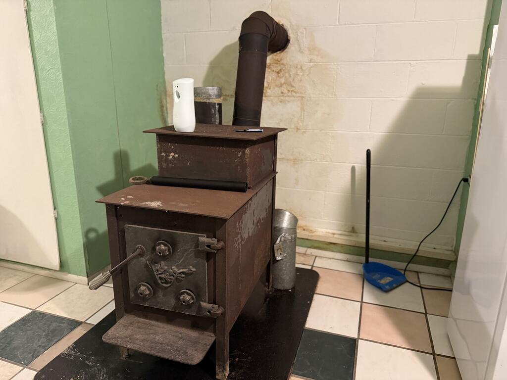 Freestanding wood-burning stove with stovepipe against a block wall, on a tiled floor.