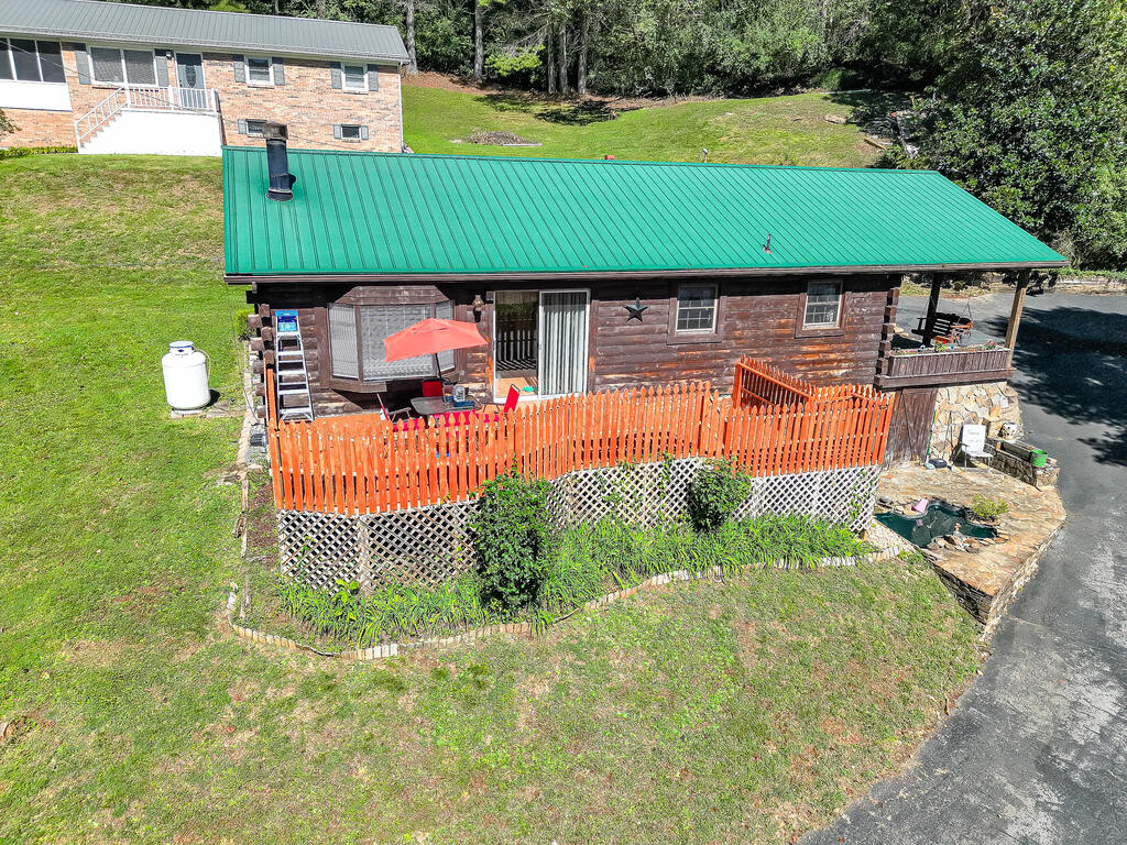 Log cabin with a green metal roof and fenced patio, viewed from above; a small pond sits beside the driveway.