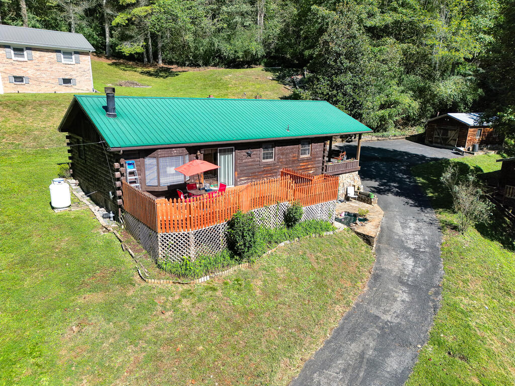 Log cabin with a green metal roof, orange-railed deck, and paved driveway in a wooded yard.
