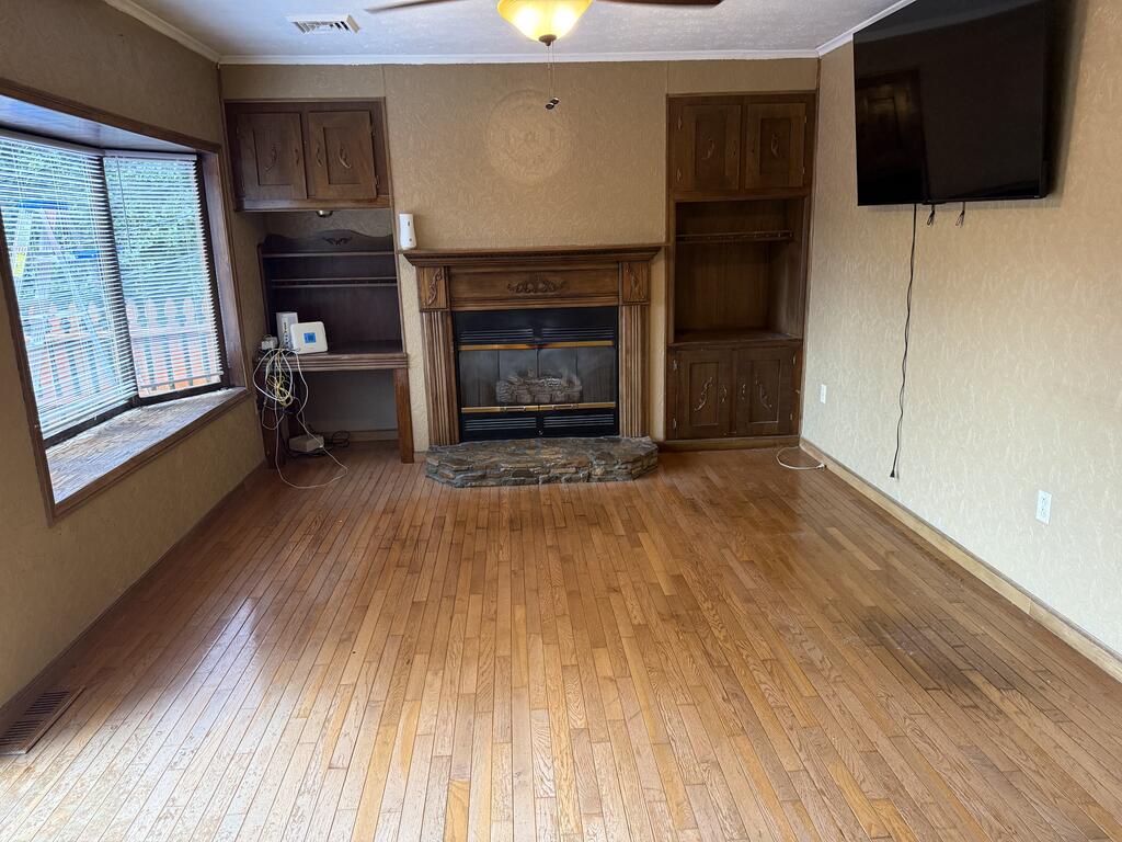 Living room with hardwood floors, a stone-front fireplace, dark wood built-ins, and a large window with blinds.