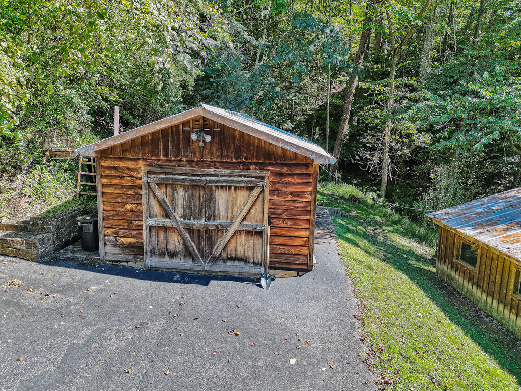 Weathered wooden outbuilding with sliding barn doors beside a paved driveway and wooded surroundings.