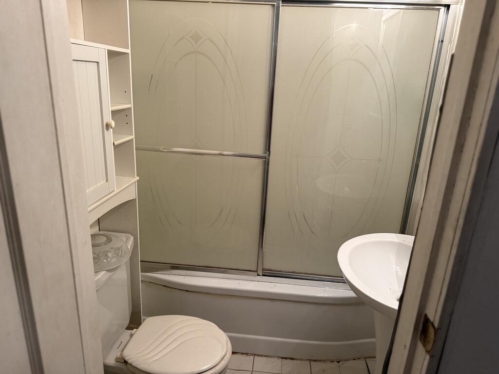 Compact bathroom with a tub, patterned sliding shower doors, toilet, and built-in shelving.