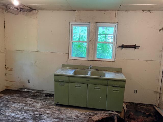 Empty room with a green sink cabinet beneath two windows and a severely damaged floor.
