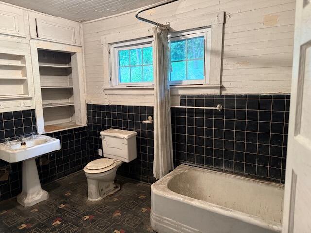 Older bathroom with black tiled walls, a pedestal sink, toilet, and bathtub beneath two windows.