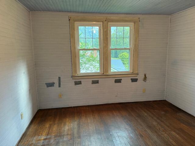 Empty room with dark hardwood floors, white paneled walls, and two windows facing greenery.