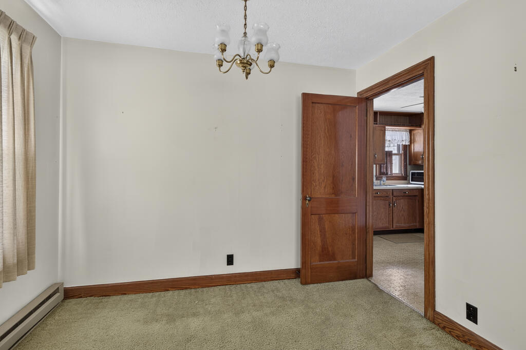 Empty carpeted room with a brass chandelier and wood-trimmed doorway to the kitchen.
