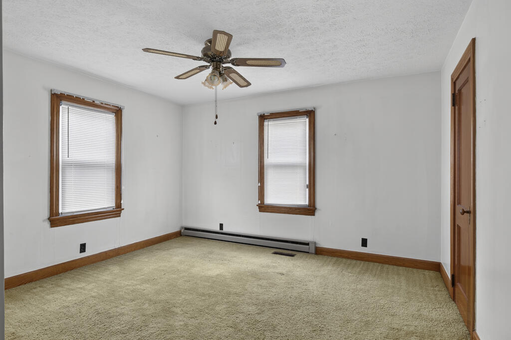 Empty bedroom with beige carpet, two wood-trimmed windows, and a ceiling fan.