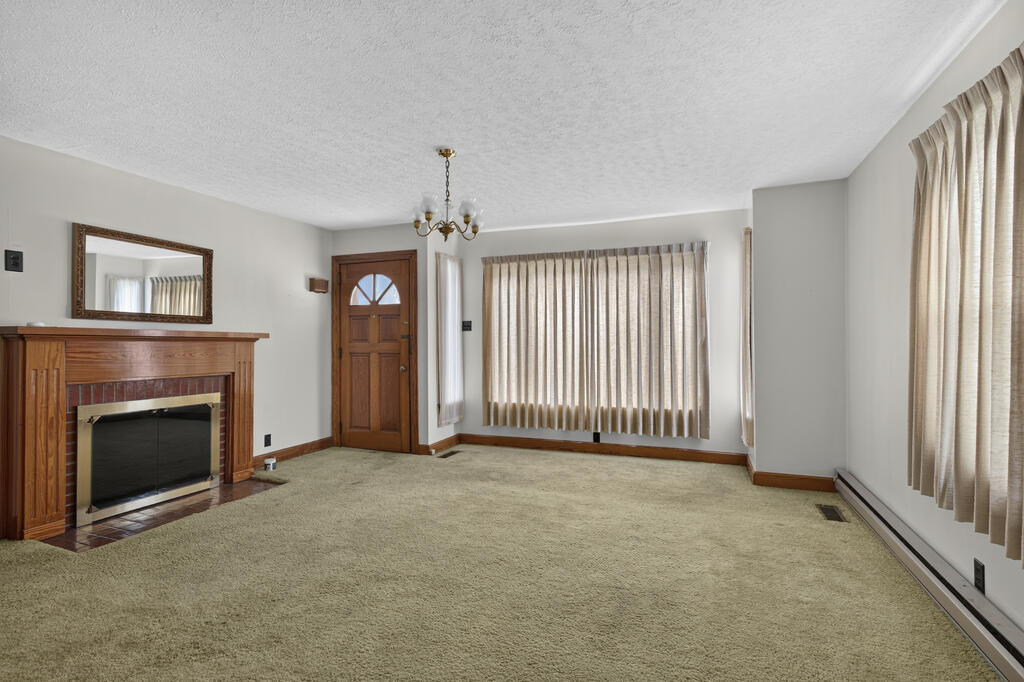 Empty living room with beige carpet, a wooden fireplace mantel, and tall curtained windows.