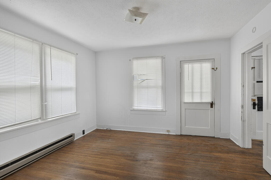 Empty room with hardwood floors, wide windows with blinds, and a windowed door.