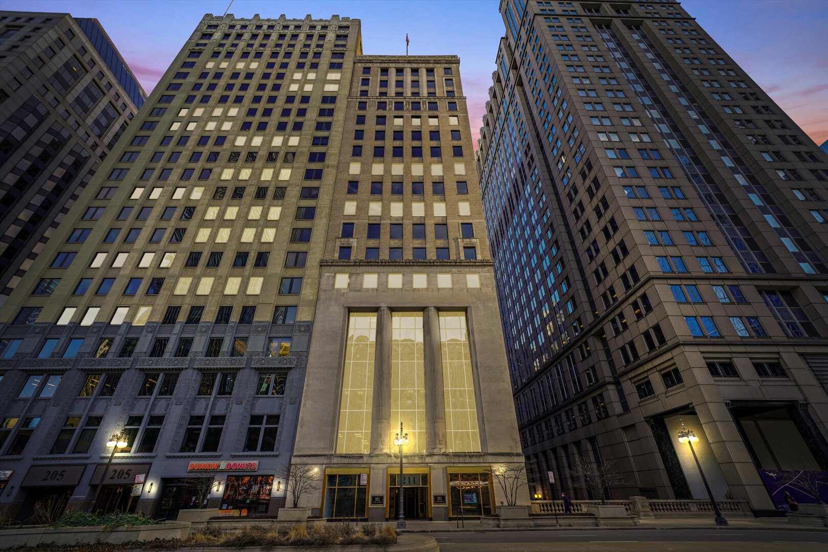Downtown office buildings at dusk, with a columned stone façade and illuminated entrance marked 211.