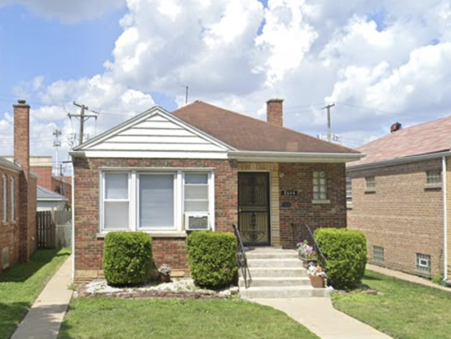 Single-story brick home with a white gable, front steps, security door, and neatly trimmed shrubs.