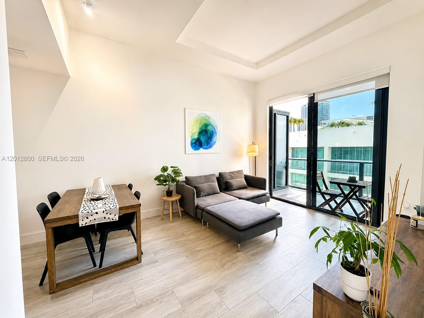 Furnished condo living room with gray sectional, dining table, and balcony doors overlooking nearby buildings.