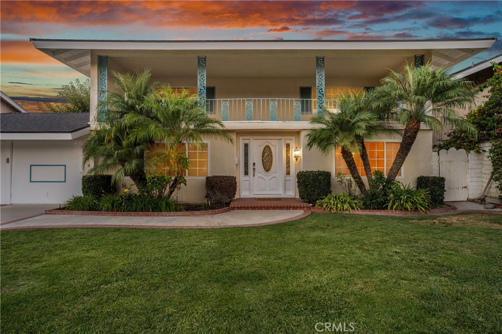 Two-story house with a broad balcony, palm trees, and a green front lawn at sunset.