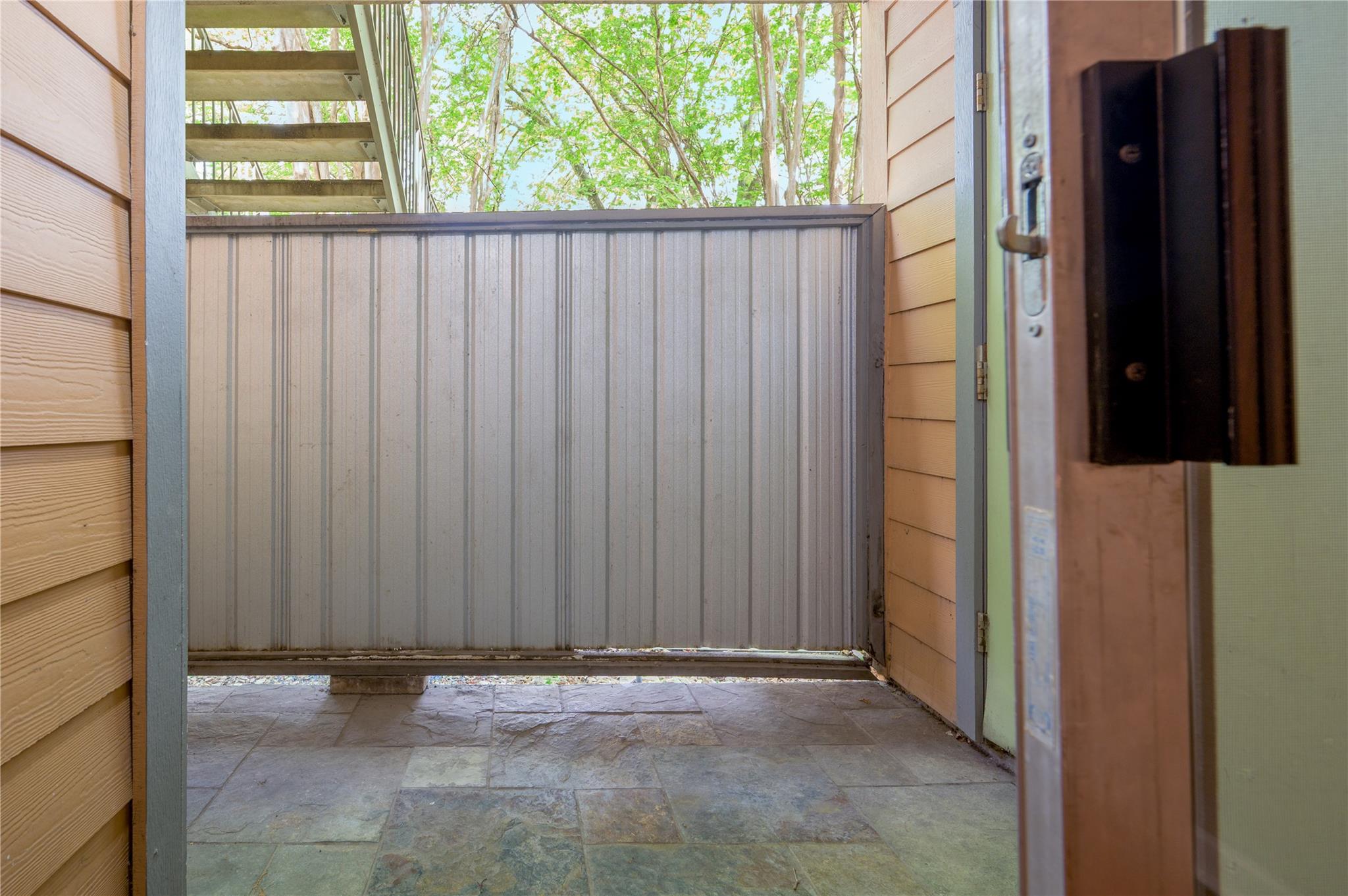 Tiled patio with a tall privacy railing, exterior stairs, and leafy trees beyond.