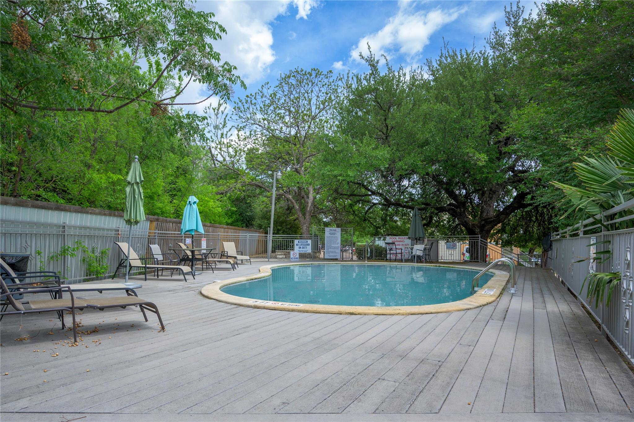 Outdoor pool with lounge chairs and umbrellas on a shaded deck beneath mature trees.