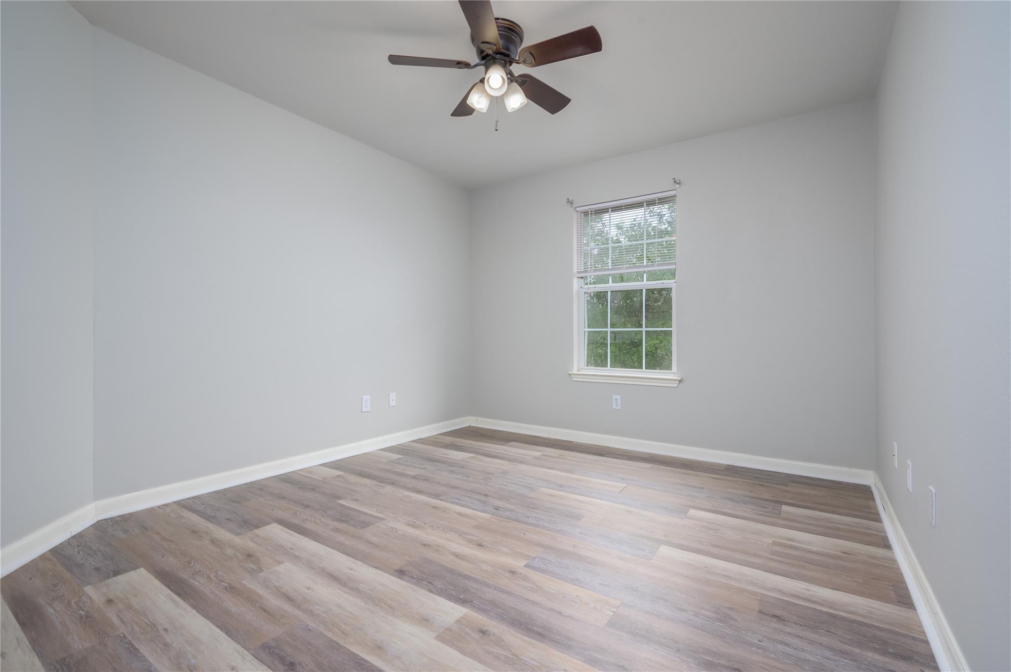 Empty bedroom with gray walls, wood-look plank flooring, a window, and ceiling fan.