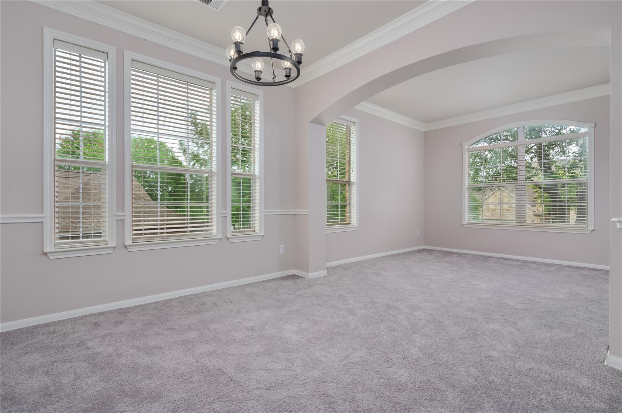 Empty carpeted room with tall windows, a wide archway, and a hanging chandelier.
