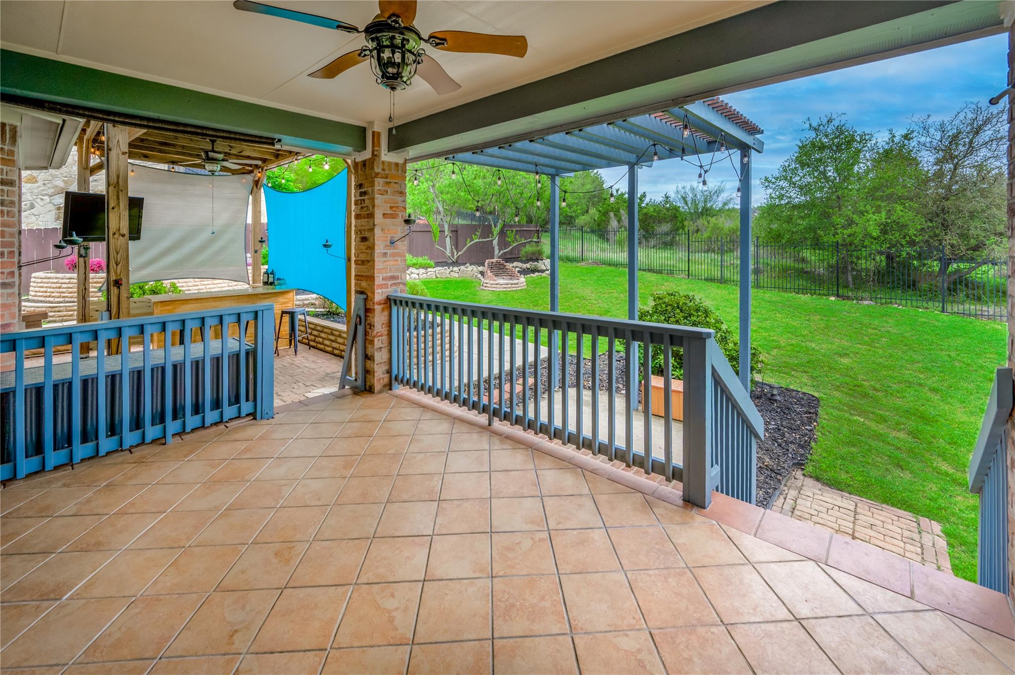 Covered patio with tiled flooring and blue railings overlooking a fenced green lawn.