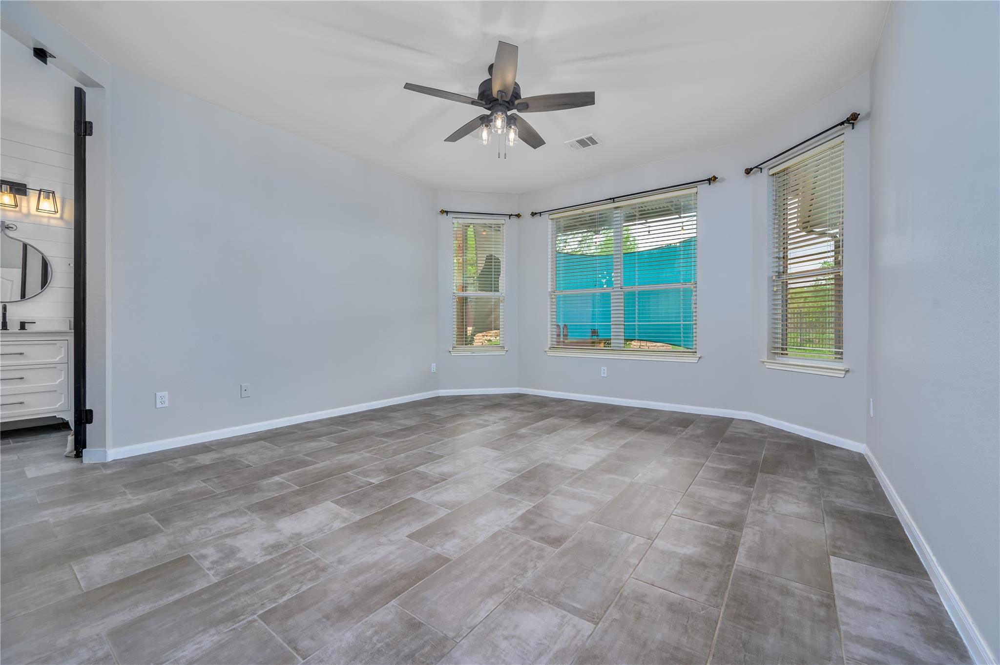 Empty room with gray tile flooring, a ceiling fan, and three windows with blinds.